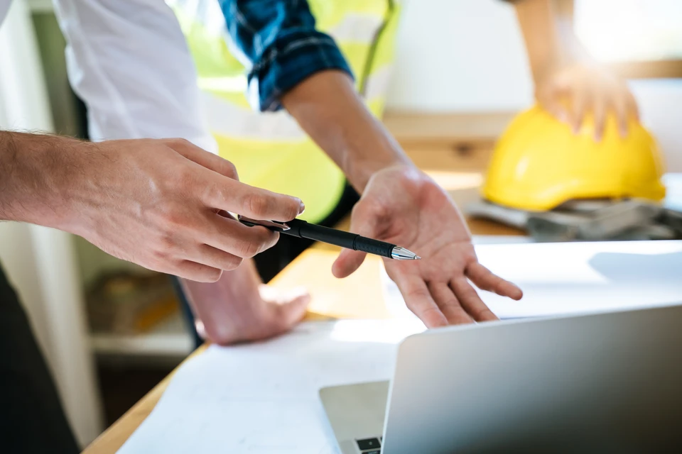 Un homme en costume travaille sur des plans architecturaux dans un bureau lumineux, tenant un stylo et des rouleaux de documents techniques